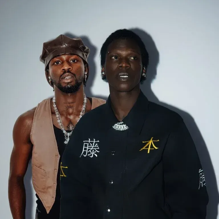 Two people posing against a plain wall, one in a brown leather hat and vest, the other in a black shirt with gold lettering.