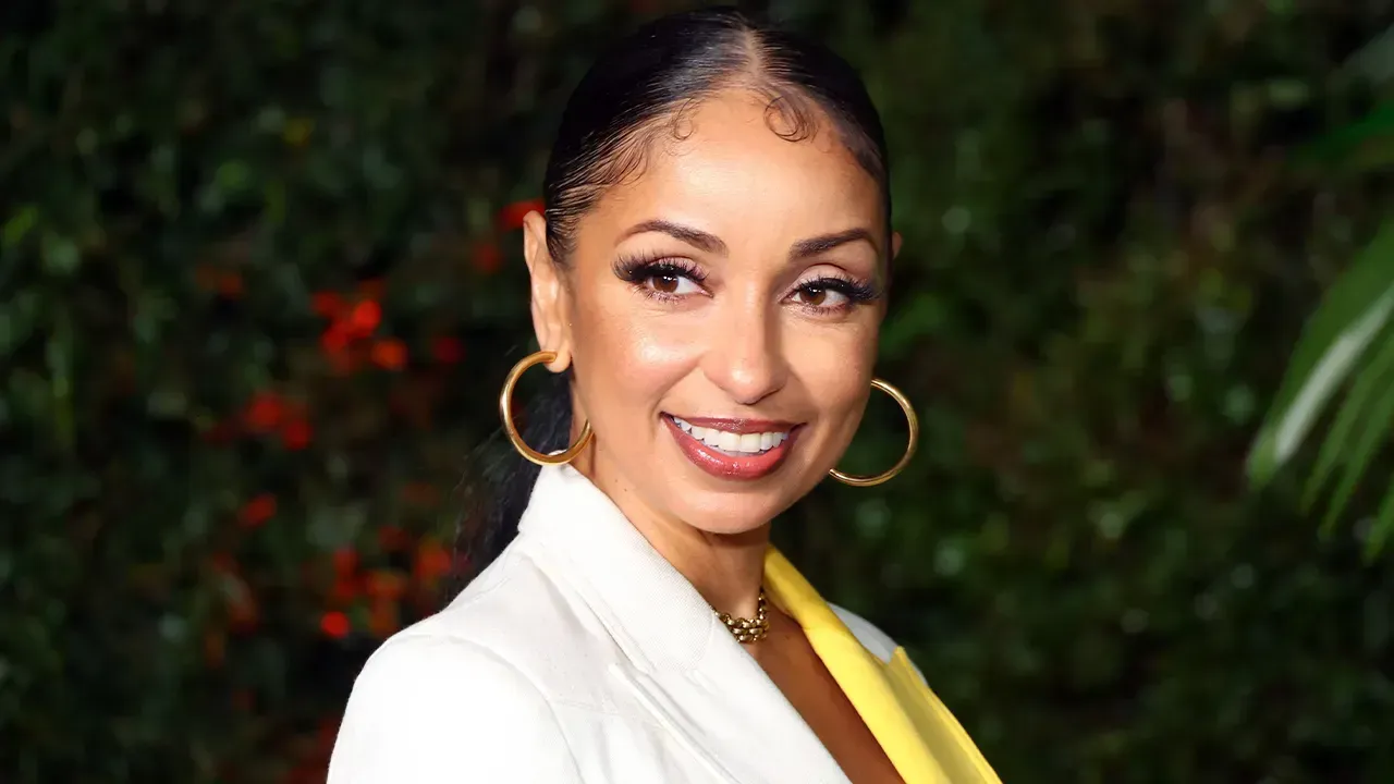 Smiling woman in a white blazer with gold hoop earrings against a leafy background