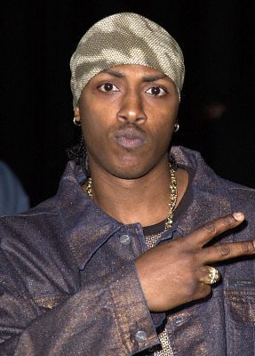Person wearing a silver headwrap and dark jacket, making a peace sign against a dark background