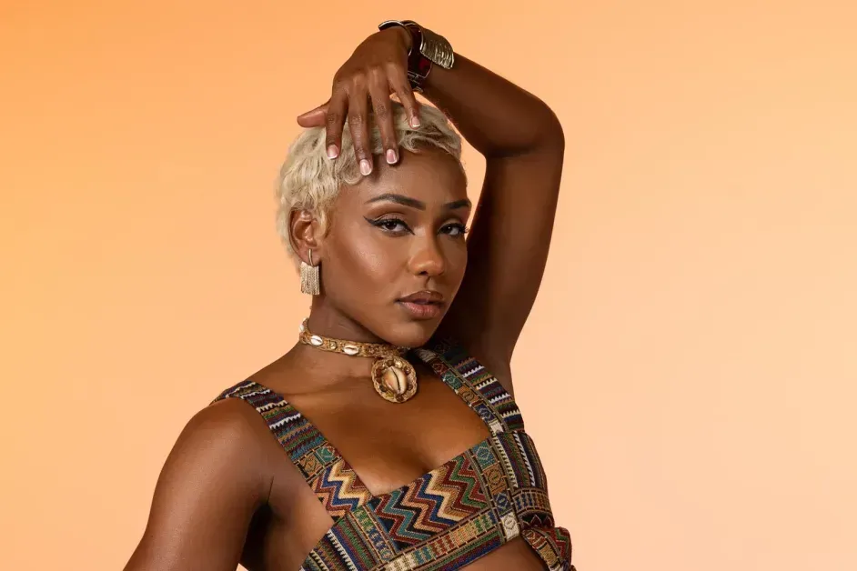 Portrait of a person posing with one arm raised, wearing patterned clothing and gold jewelry against a peach background
