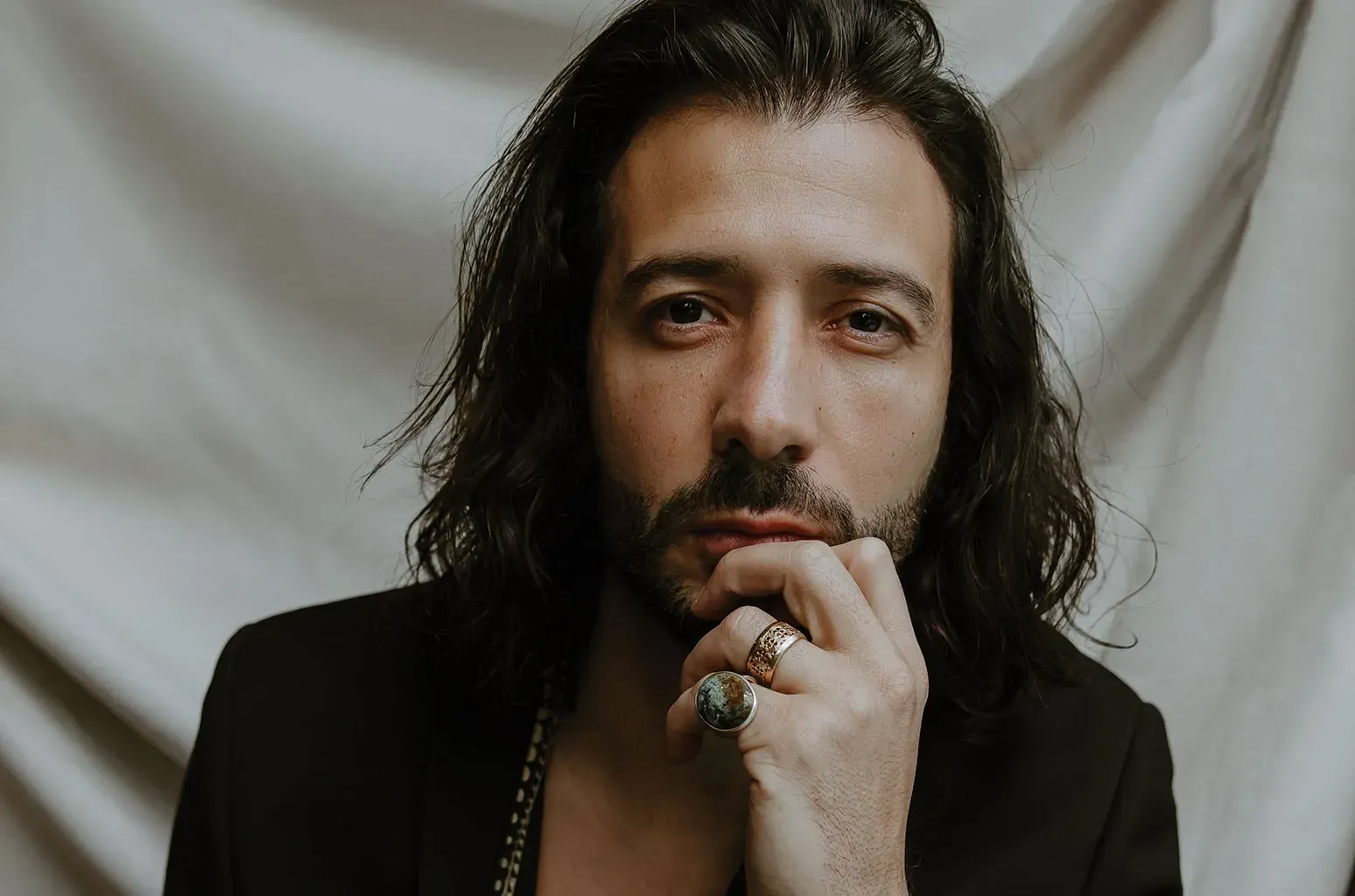Man with long dark hair in a black jacket, resting his hand on his chin against a draped backdrop