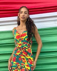Woman in a colorful patterned dress standing before red, white, and green fabric drapes.