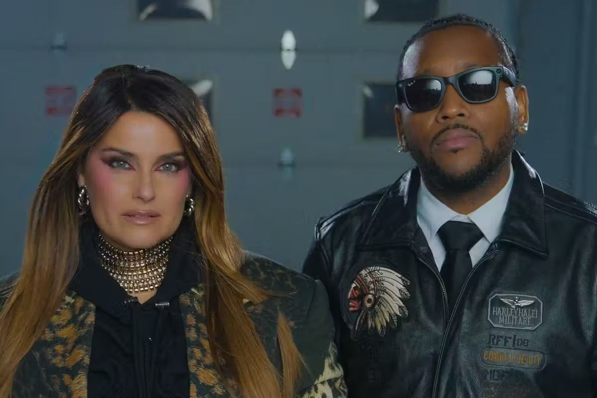 Two people in dark outfits and sunglasses stand side by side indoors, facing the camera.