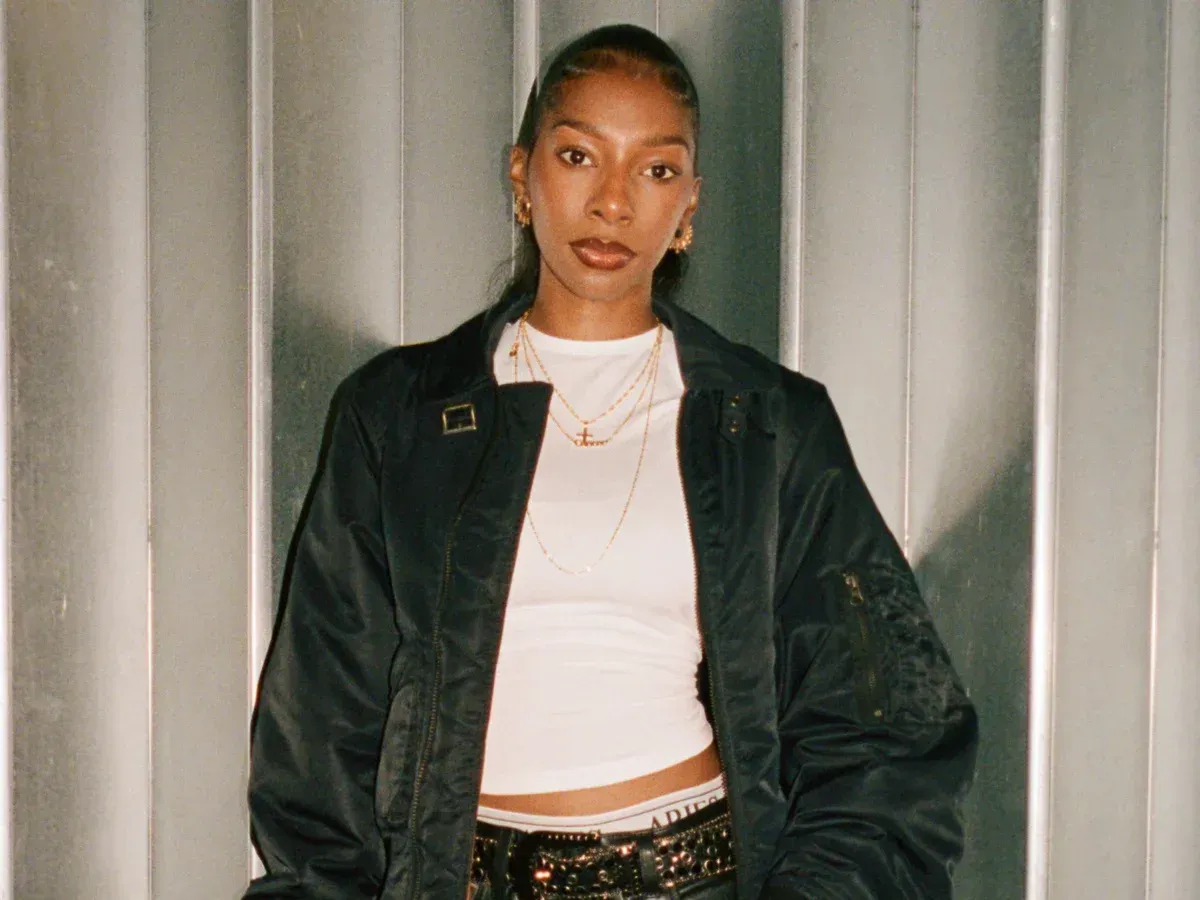 Person in a black jacket and white top standing against a corrugated metal wall.