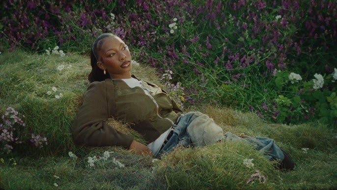 Person reclining in grass amid purple wildflowers, wearing a green jacket and jeans, with a calm expression.