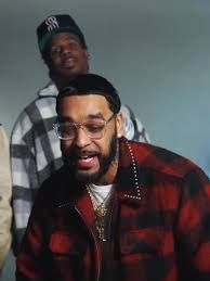Two men posing indoors, one in a red plaid jacket in front and one in a gray plaid hoodie behind.
