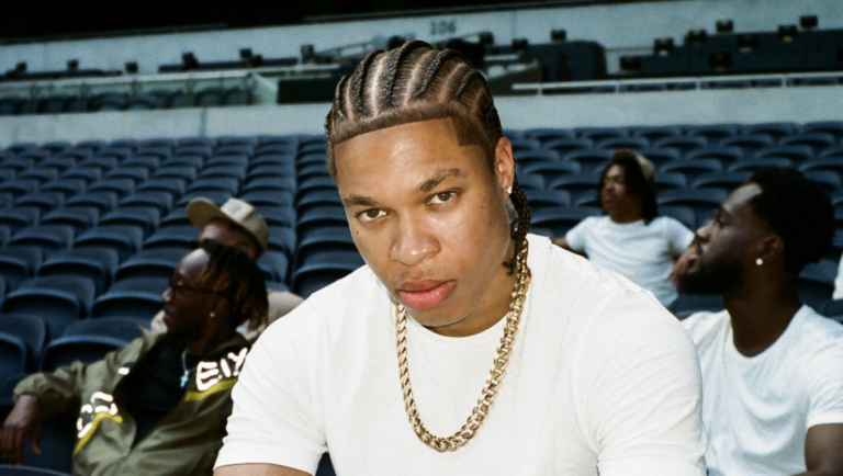 Man in white shirt and gold chain seated in an empty stadium, facing the camera with braided hair.