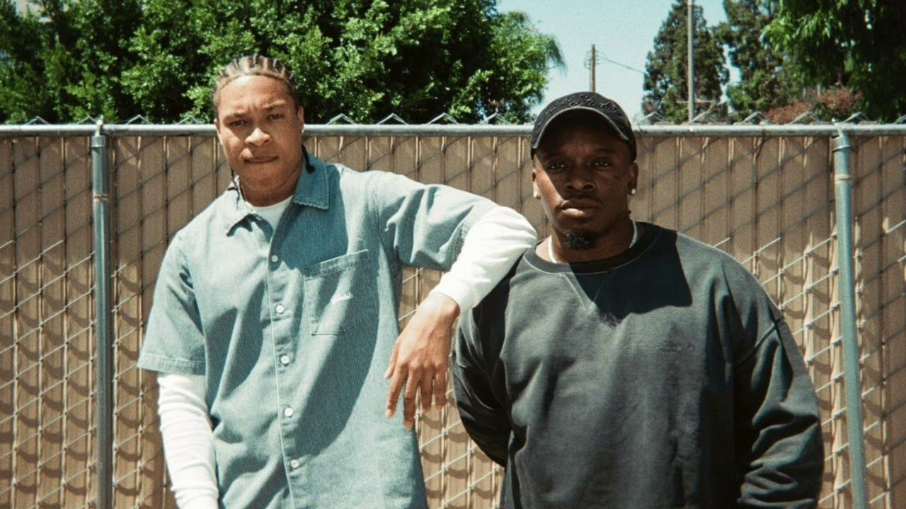 Two men stand outdoors by a wooden fence, one in a light blue shirt and one in a dark sweatshirt.