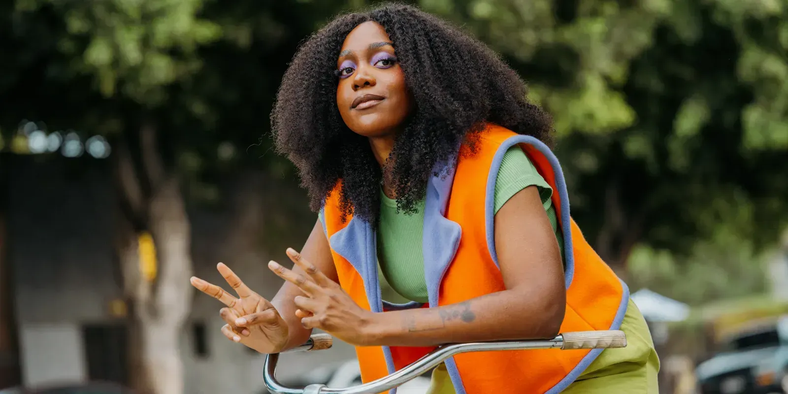 Woman in an orange safety vest gesturing outdoors with greenery in the background
