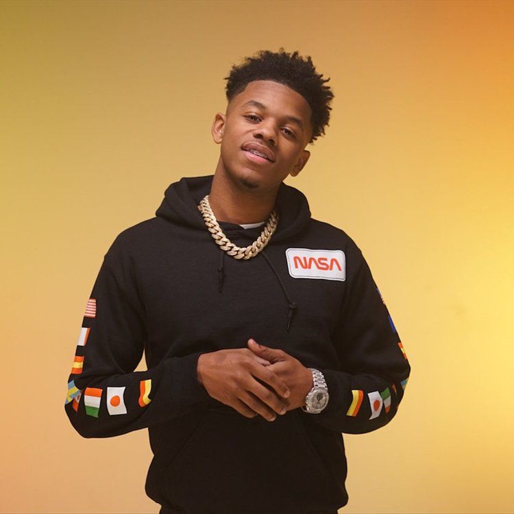 Man in black NASA hoodie and gold chain posing against a warm yellow background