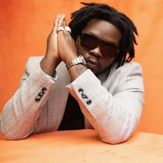 Person in sunglasses and a light blazer poses with clasped hands against an orange background.