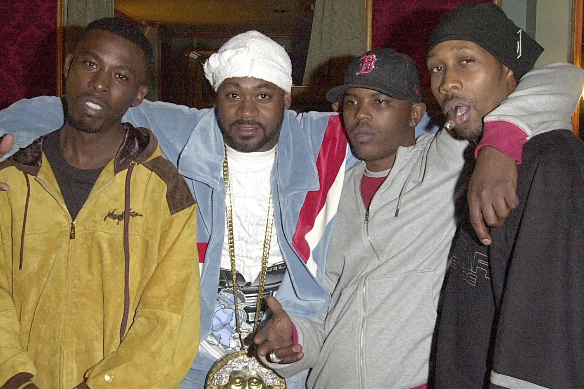 Four men stand close together indoors, wearing jackets and hats, posing for a photo.