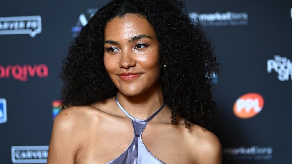 Woman with curly hair smiling at a branded event backdrop, wearing a lavender halter dress and necklace