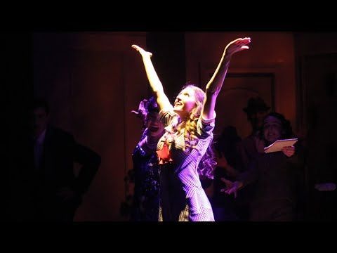 Performers on a dim stage, one in purple with arms raised under pink spotlight
