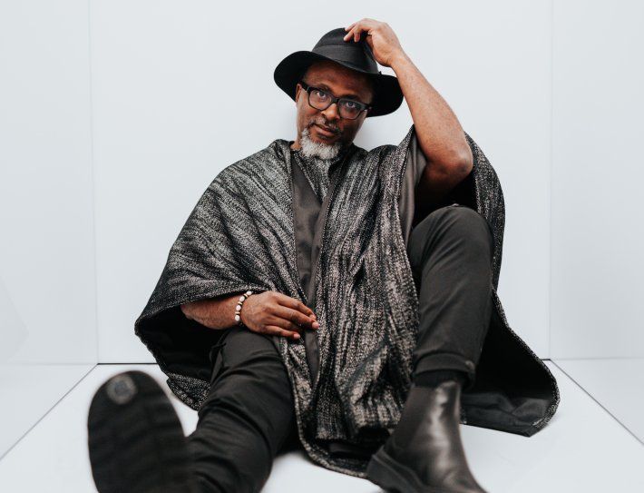 Man seated on the floor in a black hat and patterned poncho, posing against a white wall.