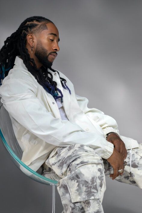 Person seated in a clear chair wearing a white jacket and patterned pants against a gray backdrop