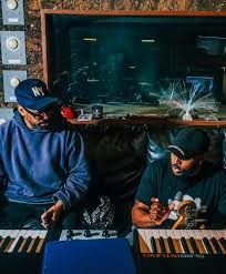 Two musicians in a studio playing keyboards, with microphones and a large window behind them