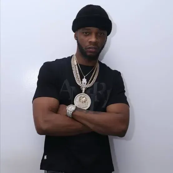 Man in black shirt and beanie standing with arms crossed, wearing layered gold chains against a plain wall.