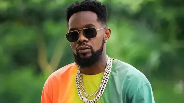 Man wearing sunglasses and gold chains in a green and orange shirt, standing outdoors against a blurred green background