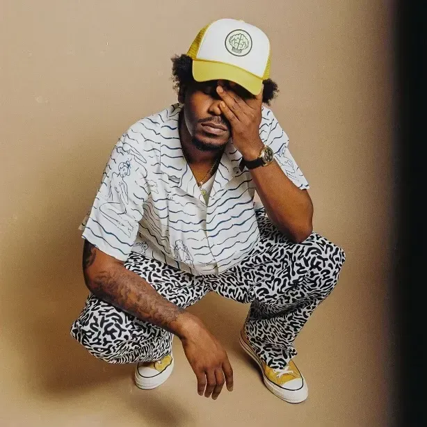 Person crouching indoors, wearing a yellow cap, patterned shirt, leopard-print pants, and white sneakers.