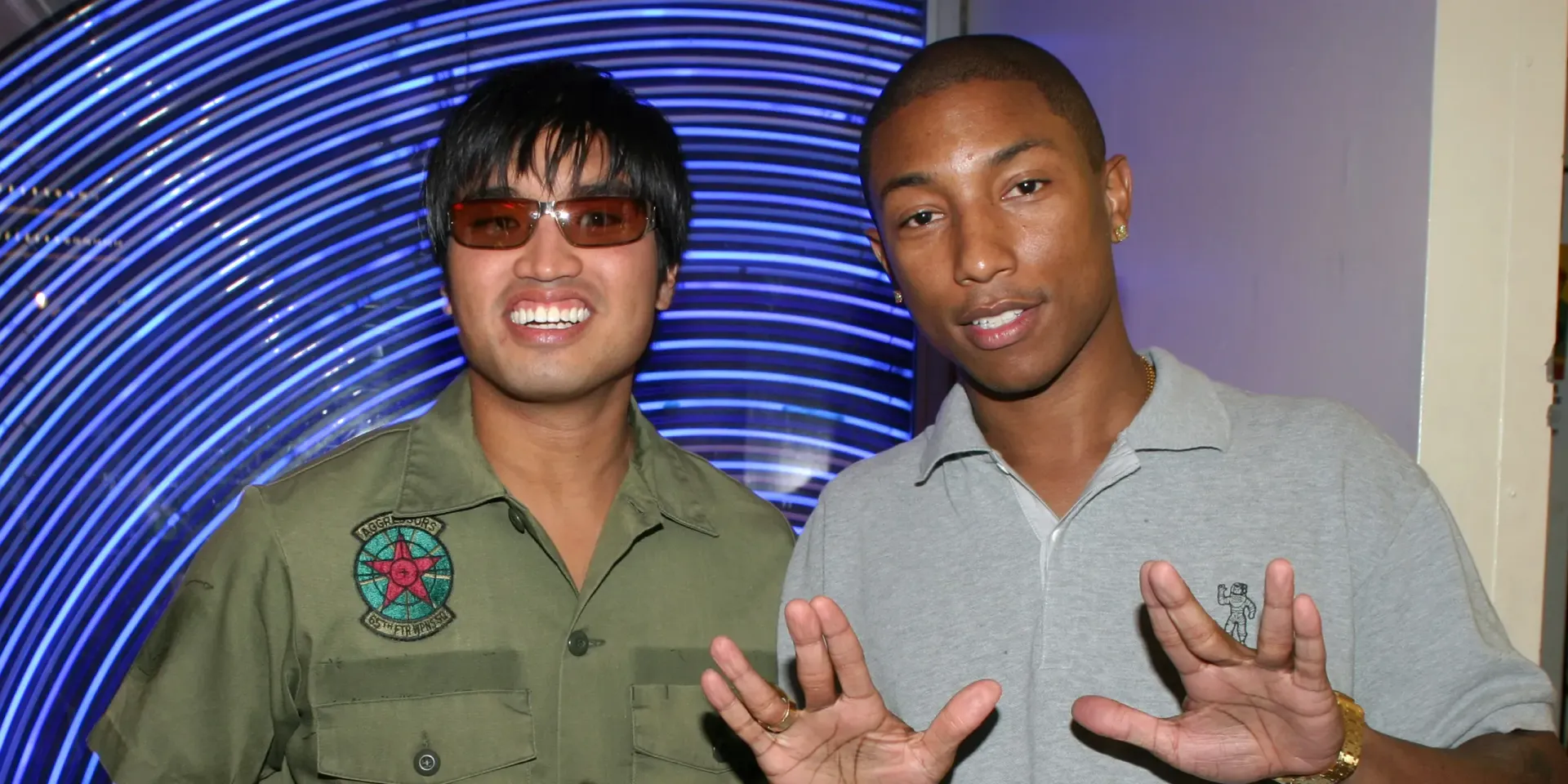 Two people posing indoors, smiling and holding up peace signs before a blue spiral backdrop