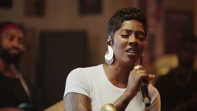 Singer holding a microphone, wearing a white shirt and large earrings, performing in a warm-lit room.