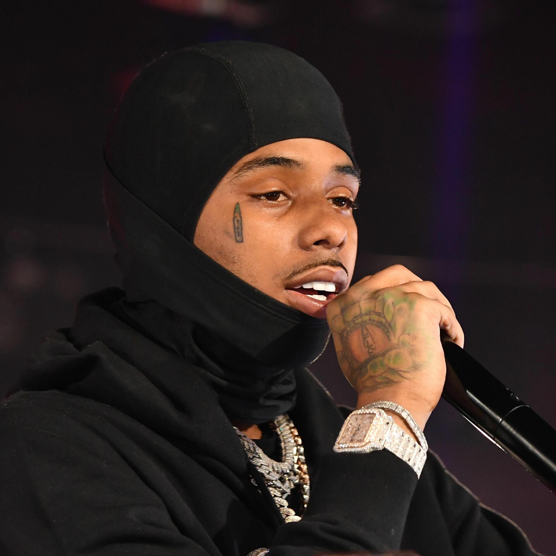 Close-up of a rapper in a black hood and mask singing into a microphone, wearing chains and a diamond watch.