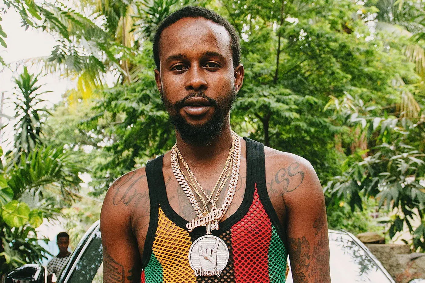 Man wearing a colorful tank top and layered necklaces, standing outdoors with greenery in the background