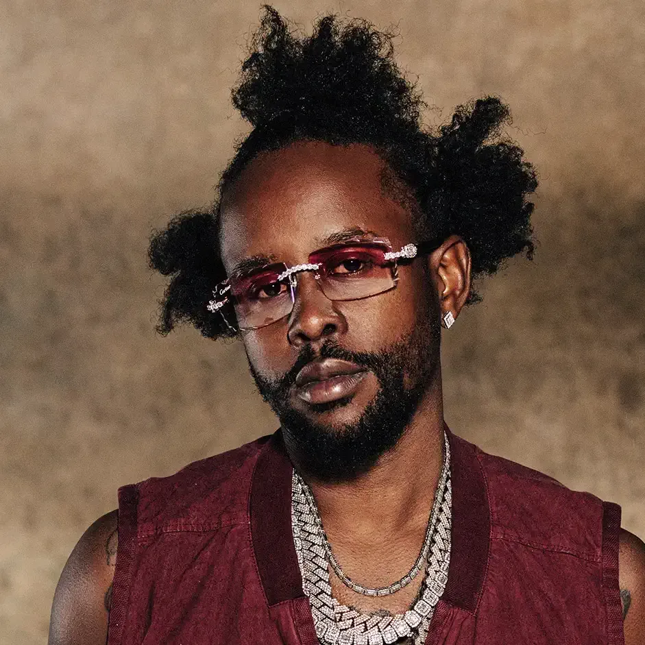 Portrait of a man with red-tinted glasses, layered necklaces, and a burgundy vest against a beige backdrop