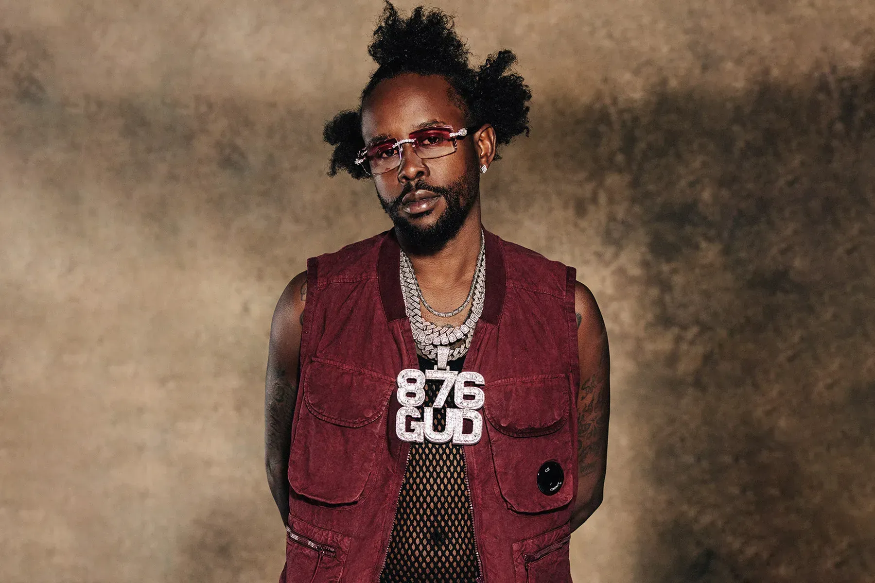 Person wearing a maroon vest and chain, standing against a textured brown backdrop.