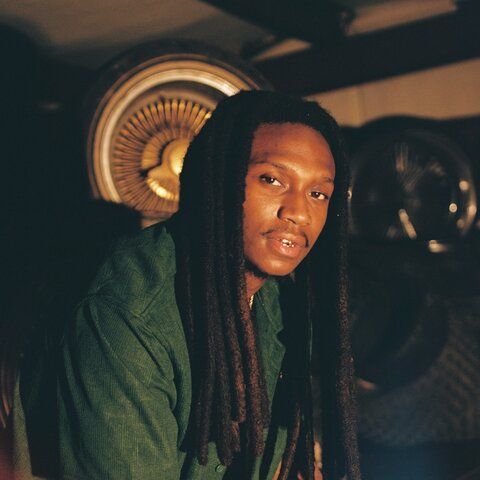 Portrait of a person with long dreadlocks in a green shirt, lit by warm light with machinery in the background