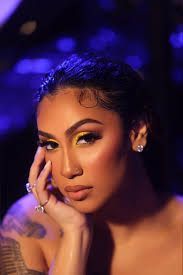Portrait of a woman with glittery makeup and curled hair, resting her face on her hand against a purple-lit background