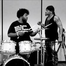 Two musicians in a black-and-white photo, one on drums and one holding drumsticks, facing each other.