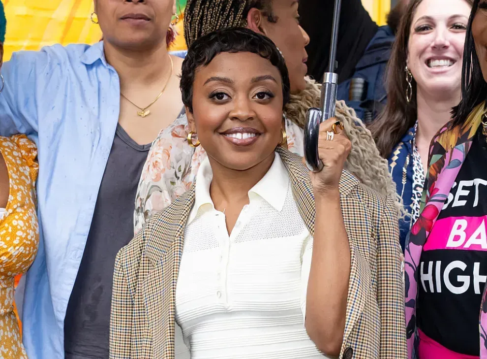 Smiling woman in a white shirt and patterned jacket stands in a crowd holding a pistol