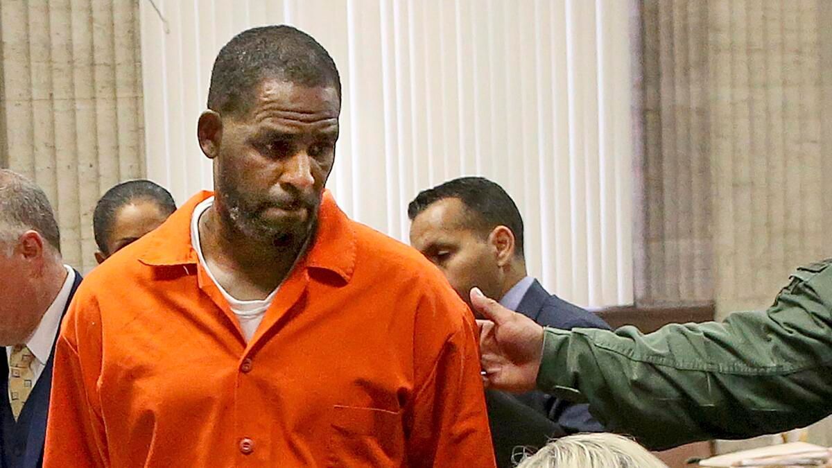 Man in orange jail uniform standing in a courtroom, looking down while another person points nearby