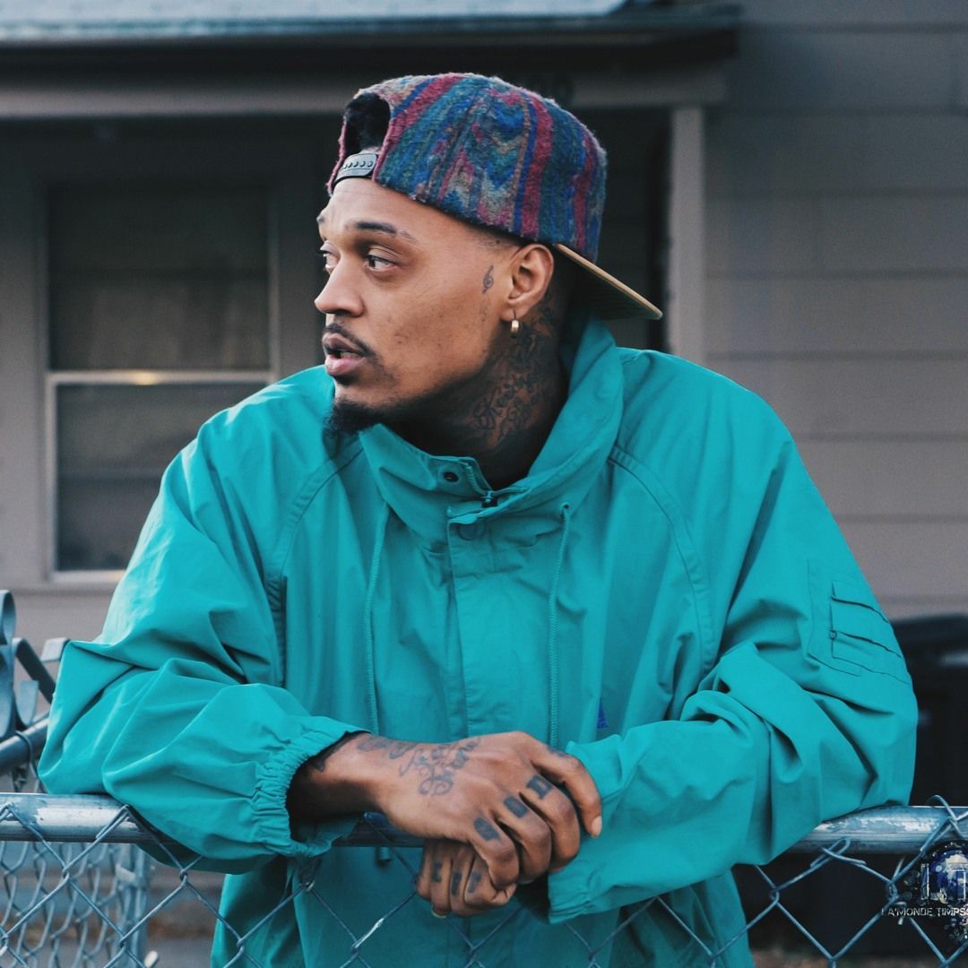 Person in a teal jacket and plaid cap leaning on a chain-link fence outdoors.