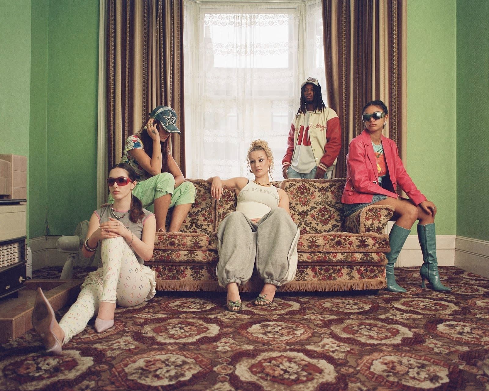 Five people posed on a patterned staircase in a vintage room, wearing colorful retro outfits and sunglasses.