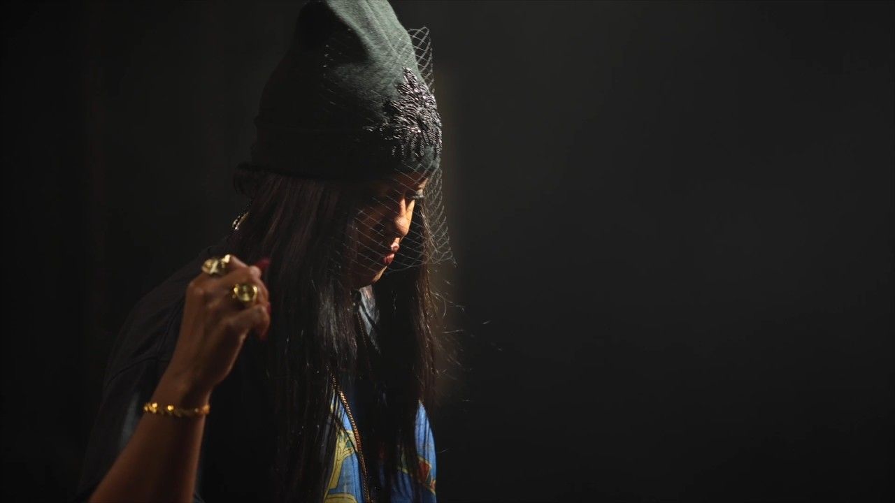 Person in a dark hat and blue top, lit dramatically against a black background.