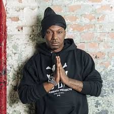 Man in black hoodie and beanie, hands pressed together, standing against a brick wall.
