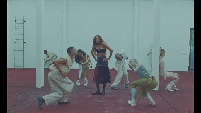 Group dancing in a studio, with a woman in dark dress centered among kneeling dancers on a red floor.