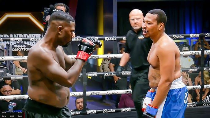 Two boxers face off in a ring, one in black gloves and one in blue gloves, with a referee behind them.