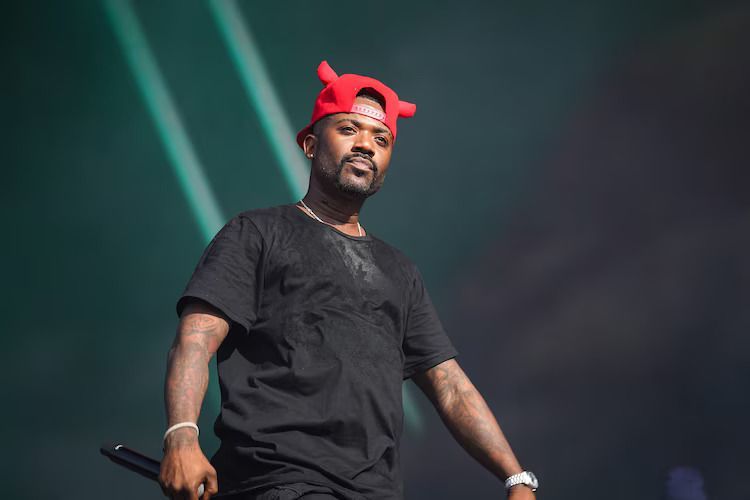 A man in a black T-shirt wearing a red bandana stands on a stage against a dark background.