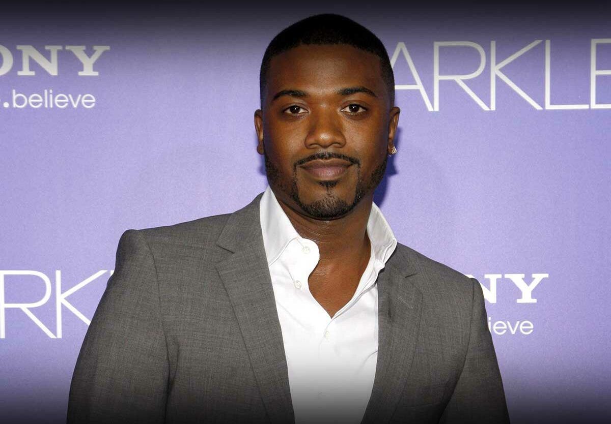 Man posing in a gray blazer and white shirt at a purple-backed red carpet event