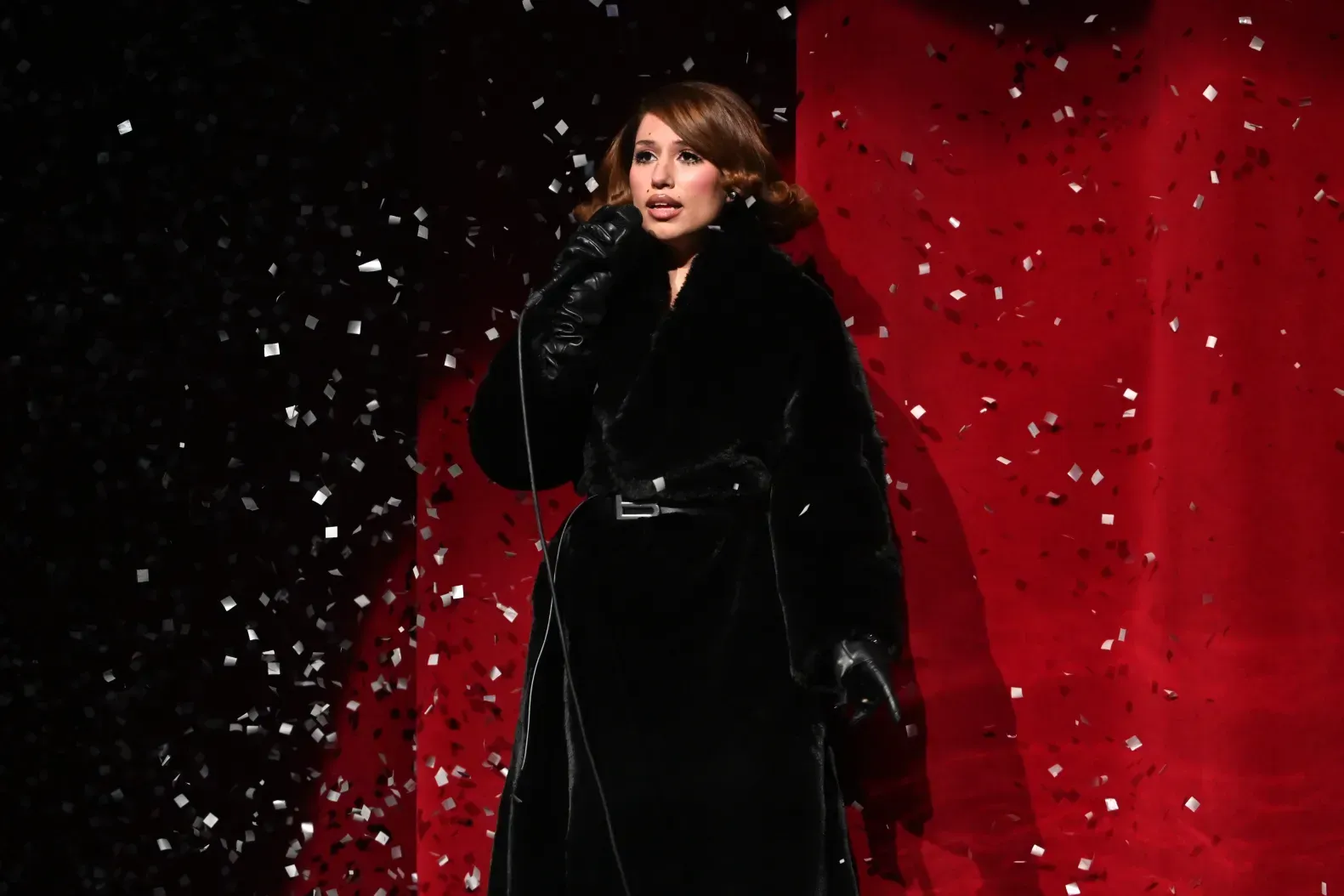 Singer in black coat performing on a red-and-black stage with falling confetti