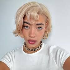 Selfie of a person with short blonde hair, gold hoop earrings, and a white shirt against a plain background