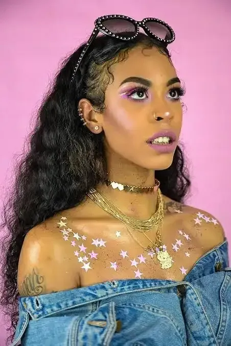 Woman with curly hair and sunglasses on head, wearing denim off-shoulder top against a pink background