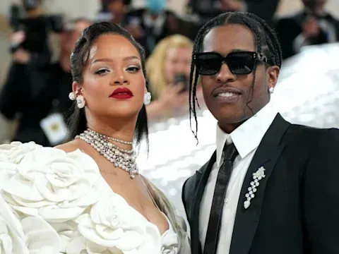 Two people posing on a red carpet, one in a white floral gown and one in a black suit with sunglasses