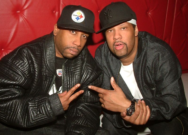 Two men in black jackets and caps posing in a red leather booth, making hand gestures.