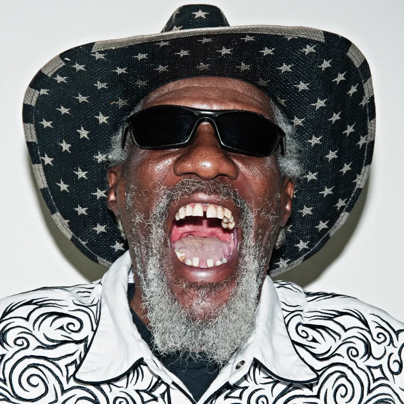Man wearing a star-patterned hat and sunglasses, shouting with mouth open against a plain background