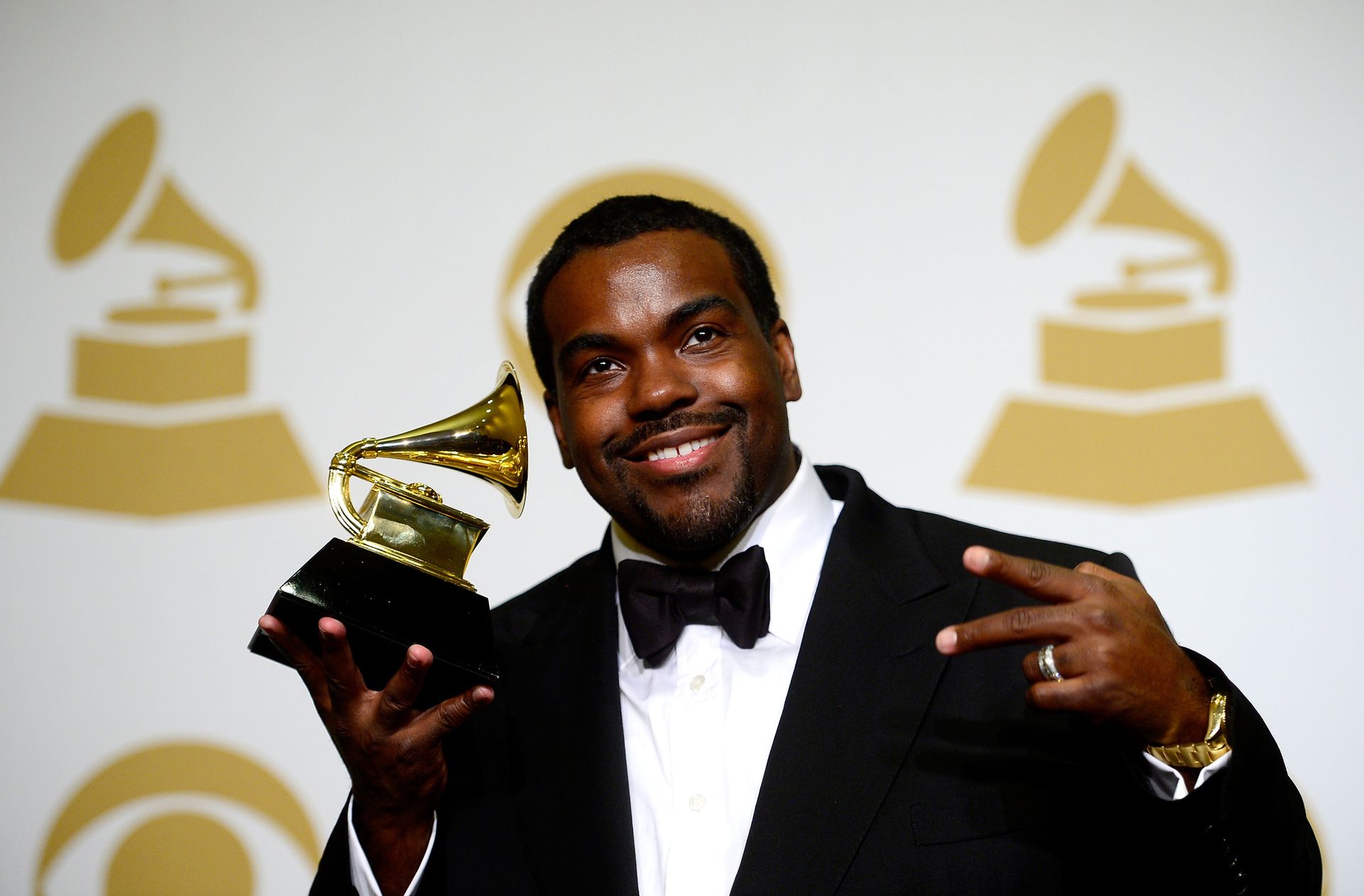 Man in tuxedo holding a Grammy trophy and smiling at a ceremony backdrop